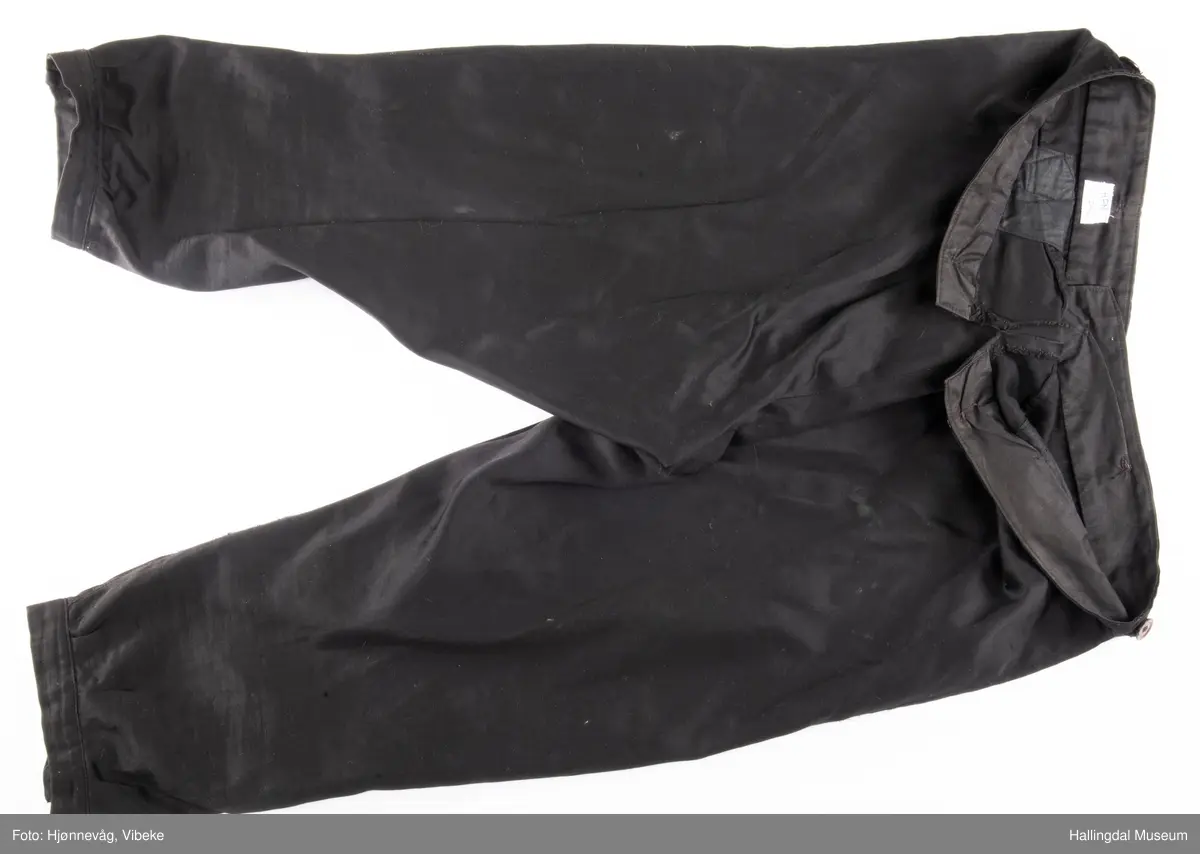 Svart knebukse i silke/bomull, glinsende stoff. 3 sølvknapper på hver fot, 4 håndsydde knapphull. Knapper i gylfen og til bukseseler på linningen. Spensel i ryggen.