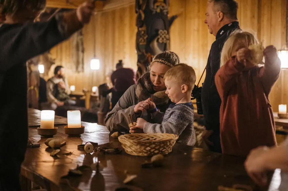 En dame og en liten gutt sitter ved et bord og lager noe av tråd. Det er levende lys på bordet og stemningsfullt med dunkel belysning. Flere mennesker sitter i bakgrunn og rundt