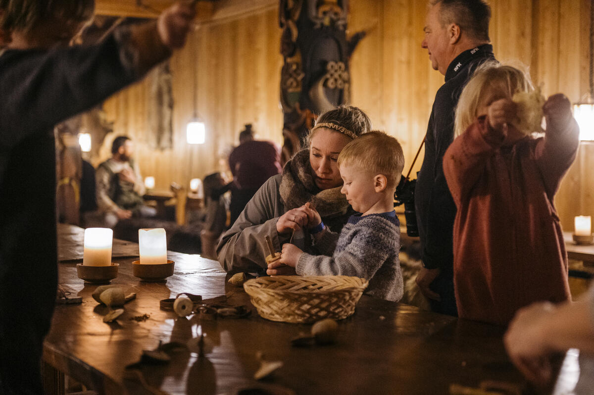 En dame og en liten gutt sitter ved et bord og lager noe av tråd. Det er levende lys på bordet og stemningsfullt med dunkel belysning. Flere mennesker sitter i bakgrunn og rundt