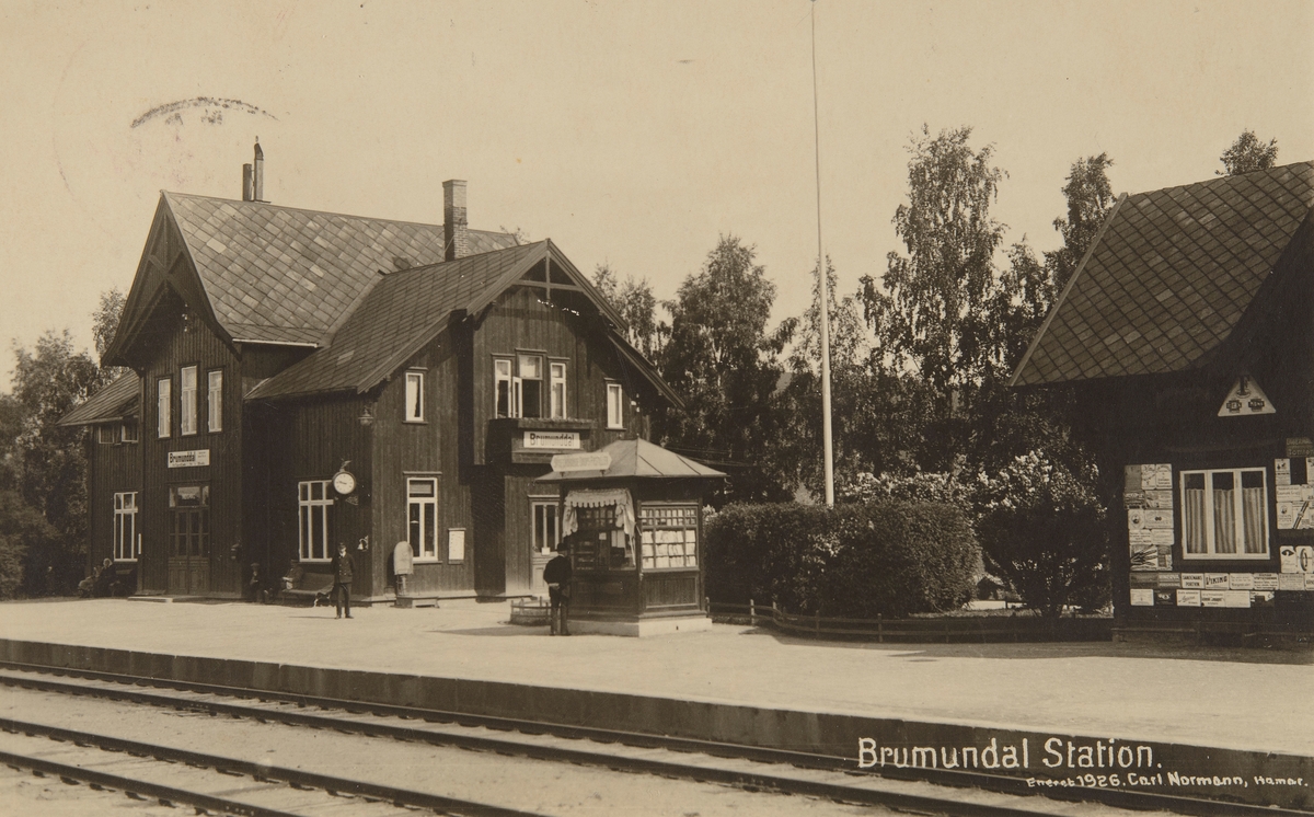 Postkort, Ringsaker, Brumunddal stasjon, Narvesen kiosk, godshus med reklame, stasjonsbetjent Sverre Ulven i uniforn, senere stasjonsmester til 1965.