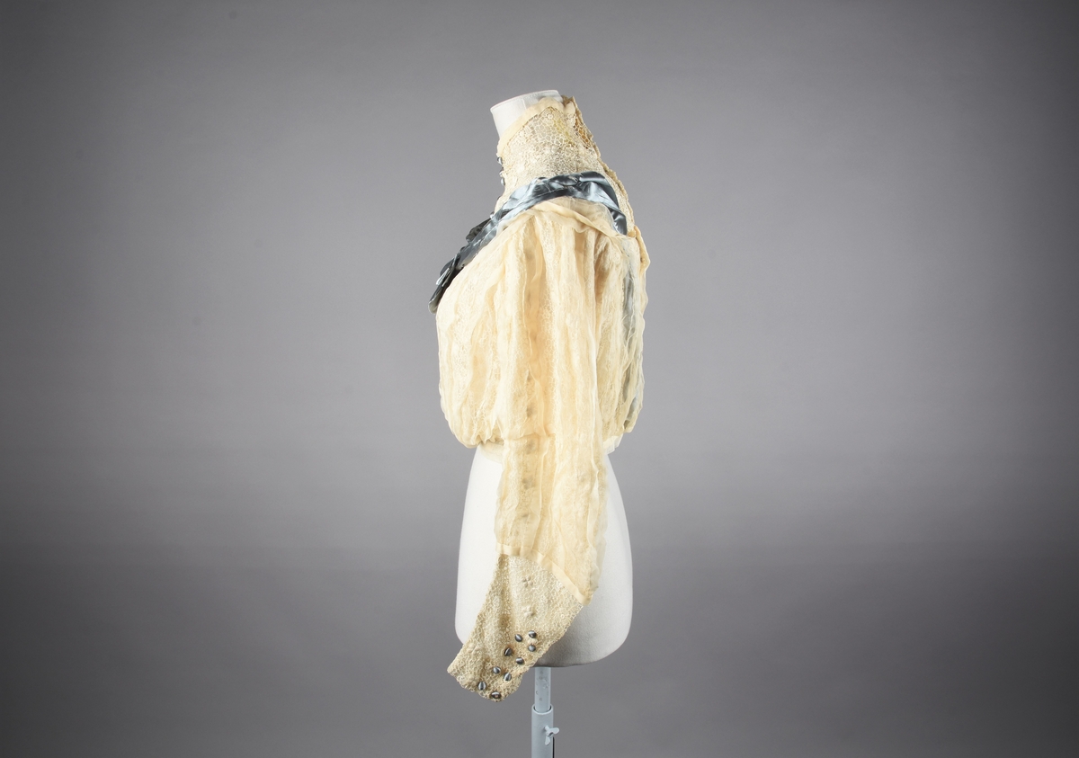 Lys elfenbensfarget bluse med tåkeblå silkebånd og silkeknapper. Den lukkes i ryggen med hekter og trådhemper. Poset front, og lange smale ermer som hektes ved håndleddene. Blusen er i guipureknipling over tyllsfór, men den høye guipure-halsen er ufóret og har tre klare gule (gulnede?) stivere. En "overbluse" av chiffon, med stor båtringning og 3/4 ermer er helt stripesydd loddrett med skiftevis bieser og 1 cm brede legg. Blusen og overblusen er sydd til livlinning av tyll. I ryggen, mellom guipuren og chiffonen, trekkes det fra linningen og over skuldrene to 8,5 cm brede silkebånd, som foran føres langs overblusens ringning ned til en sløyfe fastsydd midt foran. Atten små silketrukne og blå pynteknapper i to rekker ned på forstykket, og på hver arm ni knapper i to rekker.