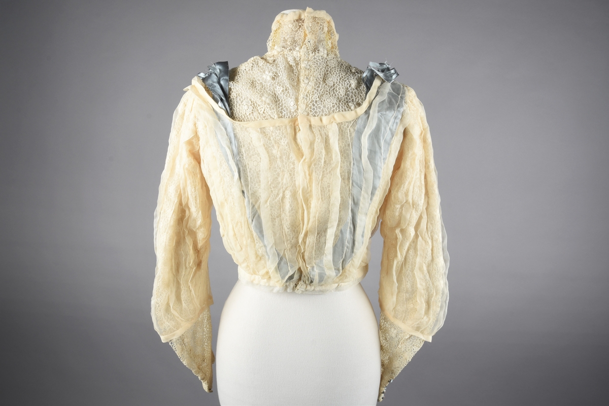 Lys elfenbensfarget bluse med tåkeblå silkebånd og silkeknapper. Den lukkes i ryggen med hekter og trådhemper. Poset front, og lange smale ermer som hektes ved håndleddene. Blusen er i guipureknipling over tyllsfór, men den høye guipure-halsen er ufóret og har tre klare gule (gulnede?) stivere. En "overbluse" av chiffon, med stor båtringning og 3/4 ermer er helt stripesydd loddrett med skiftevis bieser og 1 cm brede legg. Blusen og overblusen er sydd til livlinning av tyll. I ryggen, mellom guipuren og chiffonen, trekkes det fra linningen og over skuldrene to 8,5 cm brede silkebånd, som foran føres langs overblusens ringning ned til en sløyfe fastsydd midt foran. Atten små silketrukne og blå pynteknapper i to rekker ned på forstykket, og på hver arm ni knapper i to rekker.