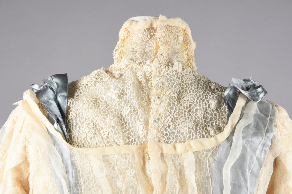 Lys elfenbensfarget bluse med tåkeblå silkebånd og silkeknapper. Den lukkes i ryggen med hekter og trådhemper. Poset front, og lange smale ermer som hektes ved håndleddene. Blusen er i guipureknipling over tyllsfór, men den høye guipure-halsen er ufóret og har tre klare gule (gulnede?) stivere. En "overbluse" av chiffon, med stor båtringning og 3/4 ermer er helt stripesydd loddrett med skiftevis bieser og 1 cm brede legg. Blusen og overblusen er sydd til livlinning av tyll. I ryggen, mellom guipuren og chiffonen, trekkes det fra linningen og over skuldrene to 8,5 cm brede silkebånd, som foran føres langs overblusens ringning ned til en sløyfe fastsydd midt foran. Atten små silketrukne og blå pynteknapper i to rekker ned på forstykket, og på hver arm ni knapper i to rekker.