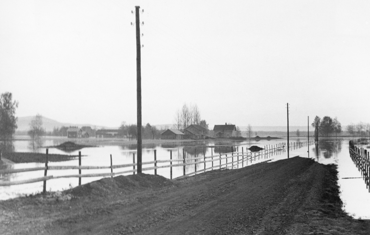 Flom på et sted som i Glomma fellesfløtingsforenings fotoarkiv er kalt Dystland i 1934. Dette er et gardsnavn i Nes kommune på Romerike, men der er avstanden til vassdraget som den nevnte organisasjonen hadde interesser i betydelig. Da dette bildet ble registrert inn i museets arkiv ble Dystland-navnet koplet til Sør-Odal kommune. Bildet er tatt fra en bygdeveg som, i likhet med omkringliggende innmark, var dekt av vann. Våningshusene på et småbruk og et litt større gardsbruk på venstre side av den nevnte vegen lå også omgitt av vann. I andre halvdel av april i 1934 begynte folk i søndre del av Hedmark og på Romerike å frykte storflom. Rapporter fra Nord-Østerdalen fortalte om vannstandsnivåer som man måtte tilbake til 1860-åra for å finne maken til, og etter kraftig regn også lengre sør på Østlandet ble flommen et problem der også. Den 19. april 1934 kom det meldinger om flomskader også på Romerike. På det stedet der dette fotografiet ble tatt var vannet såpass stillestående at skadene antakelig ble moderate.