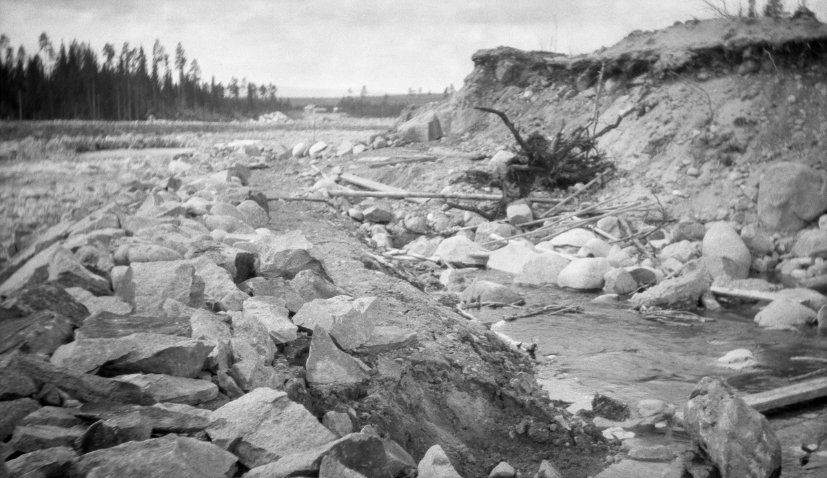 Fra den nedre delen av elva Søndre Osa i Åmot kommune i Hedmark, mellom Osfallet kraftstasjon og vegbrua der Desetvegen krysser vassdraget. Fotografiet ble tatt i 1923. Våren 1916 hadde et dambrudd i kraftverkets nordre fangdam fått elva til å grave seg et nytt, djupå og bredt løp. Erosjonseffekten var enorm, og den ble ikke mindre etter det neste store flomåret, 1920. For å forebygge ytterligere erosjonsskader fikk åmotingene støtte fra Vassdragsvesenet til å legge opp kraftige steinvoller langs elveløpets ytterkanter for å forebygge ytterligere erosjonsskader. I bakgrunen på dette fotografiet skimter vi småbruket Osmoen.