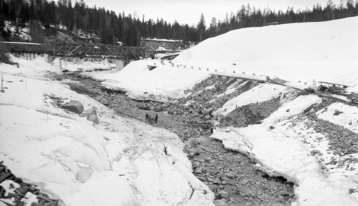 Fra det nye løpet elva Søndre Osa i Åmot i Hedmark grov seg da den nordre fangarmen for dammen ved Osfallet sviktet under flom våren 1916.  Da kraftverket skulle settes i drift igjen etter denne katastrofen måtte det etableres et nytt vannreservoar noe høyere oppe i elveløpet, og dette fotografiet er tatt mellom det som da ble kalt «Nydammen» og «Fallet». Bildet gir et glimt fra Vasdragsvesenets forbygningsprosjekt som skulle stabilisere elveløpet og de nye installasjonene som var bygd der. Nede i elvetfaret skimter vi tre arbeidere i aktivitet, og på en avsats i elveskråningen til høyre i bildet hadde man støpt ei rekke av fundamenter som antakelig skulle bære et turbinrør. I bakgrunnen var det ei fagverksbru - antakelig av tre, men på betongstøpte landkar. Etter snødekket å dømme er dette fotografiet tatt i overgangsfasen mellom vinter og vår. Dette var en ideell periode for slike arbeider, for da var vannføringa i vassdraget som regel meget beskjeden. Bildet skal være fra 1928. I mellomkrigstida var slike forbygningsarbeider i vassdragene ofte sysselsettingsprosjekter for folk som - i hvert fall på denne årstida - manglet fast inntekt.