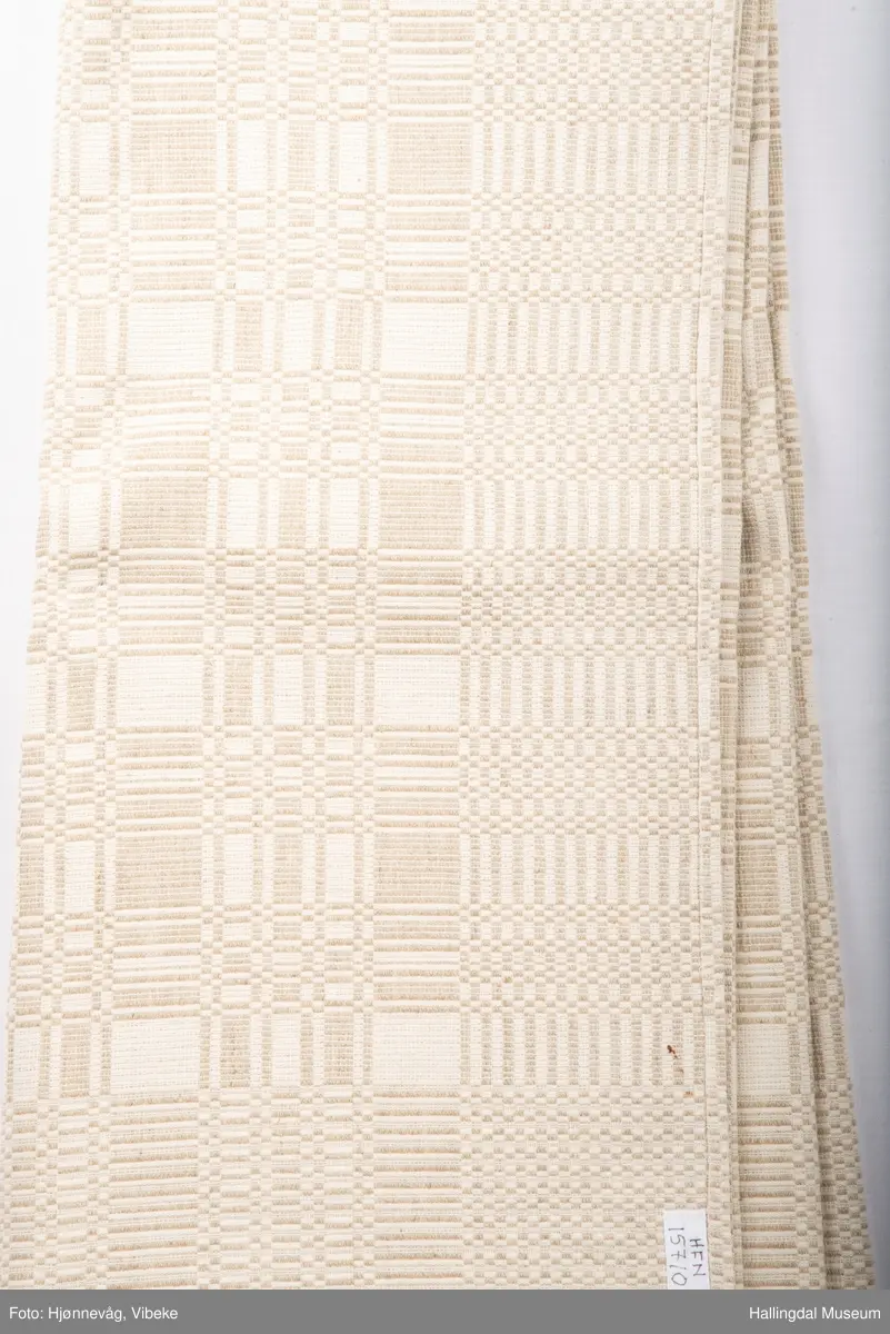 Linduk, linrenning og lin i innslag, beige. Danner ruter ig striper, tavlebragd. Duken er vevd i to lengder som er sydd sammen på midten med håndsøm.