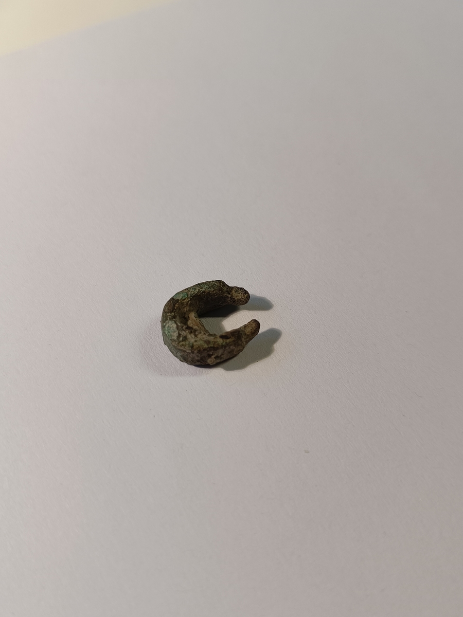 Lite, hesteskoformet beslag, uvisst til hvilket bruk.  Det går en nagle tvers gjennom buen på beslaget. I beslagets to ender, smalner sidene av til halvparten av sin øvrige bredde. beslaget har rester av forgylling. 
