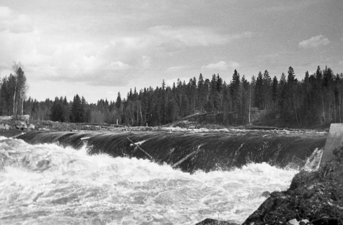 Dammen som skulle stuve opp vann til Kvernfallet kraftverk i elva Søndre Osa i Åmot kommune i Hedmark, fotografert våren 1936, like etter at anleggsarbeidet var ferdig. Her renner vannet over en avrundet terskel, utført i armert betong. Denne konstruksjonen er plassert på tvers av elveløpet, like framfor ei oppoverskrånende bergflate, som leder vannet fra nedfallssona mot den søndre elvebredden, før det føres videre vestover i det gamle elveløpet. 1936 var første fløtingssesong etter at dammen var ferdig, og det var antakelig en viss spenning knyttet til hvordan tømmeret ville komme seg over damkrona. Kraftproduksjonen fra dette anlegget startet først på høsten, fire-fem måneder etter at dette fotografiet ble tatt.