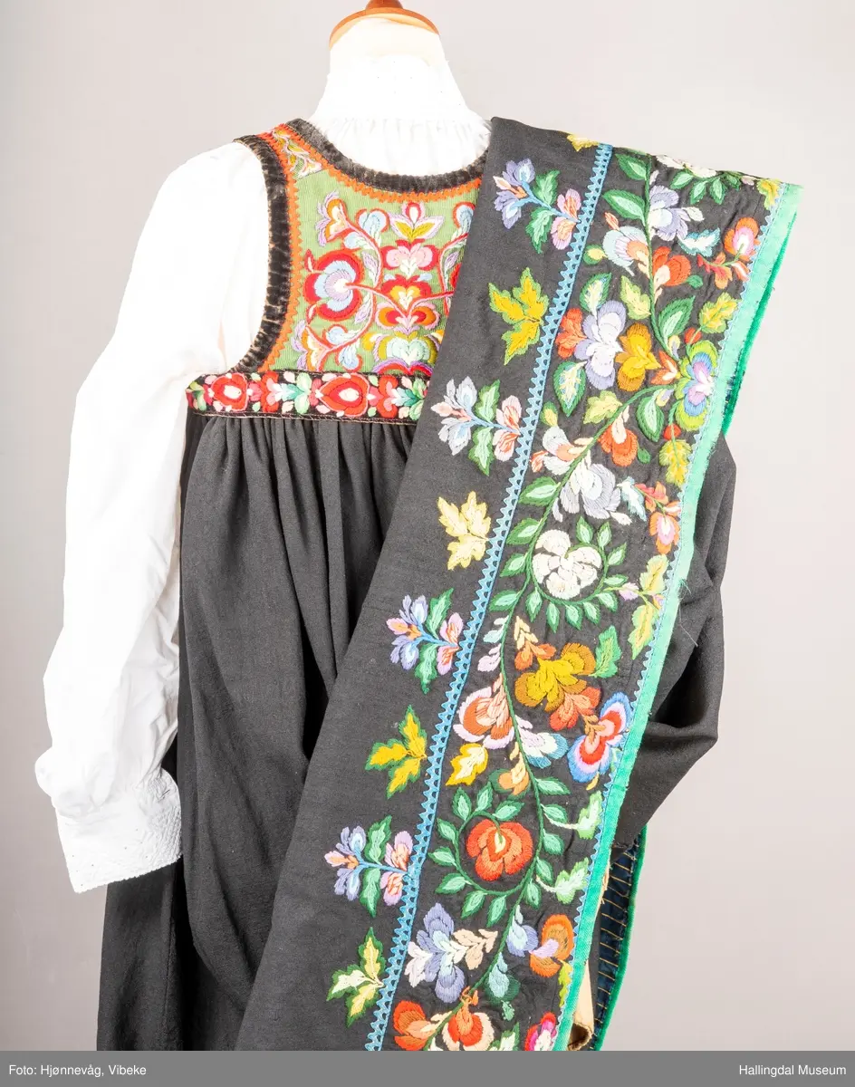 Ull, toskaft, sydd sammen av 5 høyder ca. 52 cm brede. Håndsydd. Broderi med ullgarn, mange ulike farger, akantusranke nederst. Blå kroklisse, grønn ullkant nederst. Lomme i høyre side. Blå skoning med hvite striper. Livet er grønt, ripsbinding. Svarte føyelskanter m/ rød kroklisse. Hvitt lerretsfor. Broderte hallingroser med ullgarn, sydd gjennom foret. Sølvspenner på hver side av livet med et kjede.