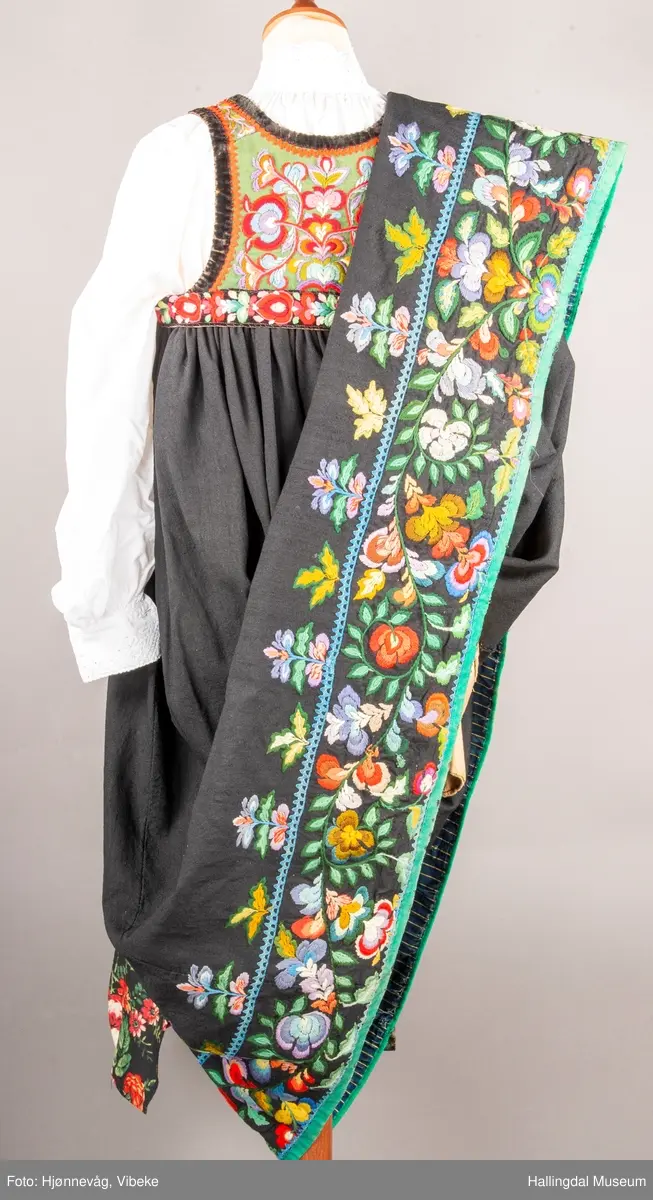 Ull, toskaft, sydd sammen av 5 høyder ca. 52 cm brede. Håndsydd. Broderi med ullgarn, mange ulike farger, akantusranke nederst. Blå kroklisse, grønn ullkant nederst. Lomme i høyre side. Blå skoning med hvite striper. Livet er grønt, ripsbinding. Svarte føyelskanter m/ rød kroklisse. Hvitt lerretsfor. Broderte hallingroser med ullgarn, sydd gjennom foret. Sølvspenner på hver side av livet med et kjede.