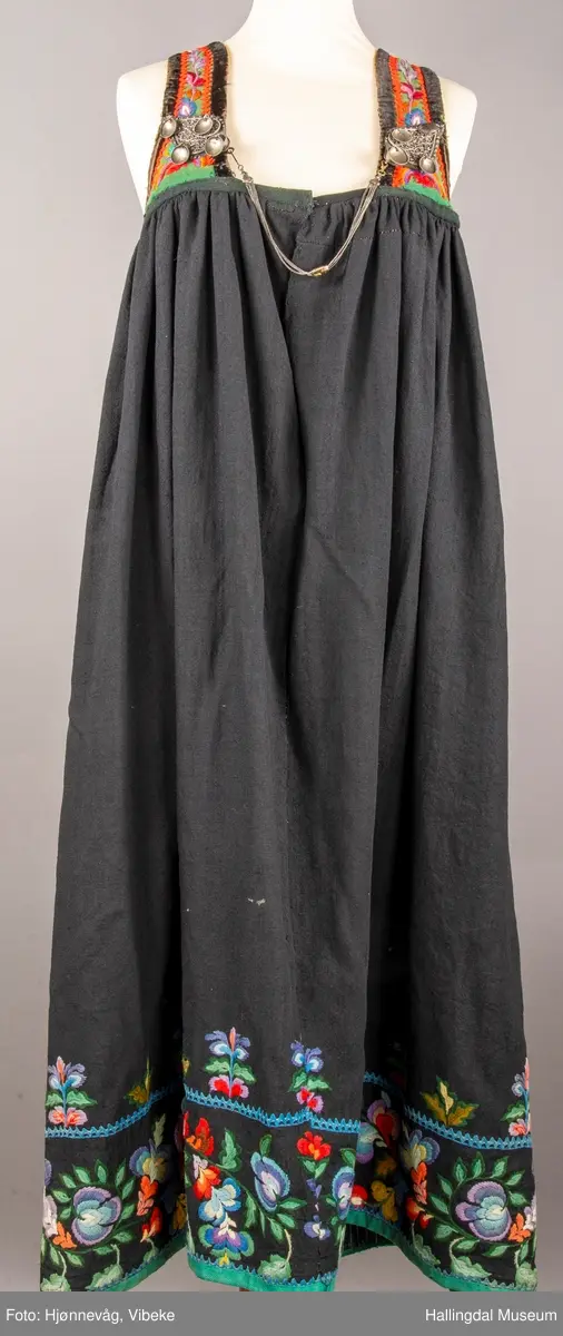 Ull, toskaft, sydd sammen av 5 høyder ca. 52 cm brede. Håndsydd. Broderi med ullgarn, mange ulike farger, akantusranke nederst. Blå kroklisse, grønn ullkant nederst. Lomme i høyre side. Blå skoning med hvite striper. Livet er grønt, ripsbinding. Svarte føyelskanter m/ rød kroklisse. Hvitt lerretsfor. Broderte hallingroser med ullgarn, sydd gjennom foret. Sølvspenner på hver side av livet med et kjede.