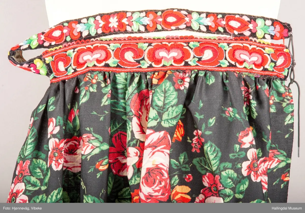 Storblomstrete ullmusselin med brodert kant øverst, hallingroser. Ryggbåndet er festet til forkle.