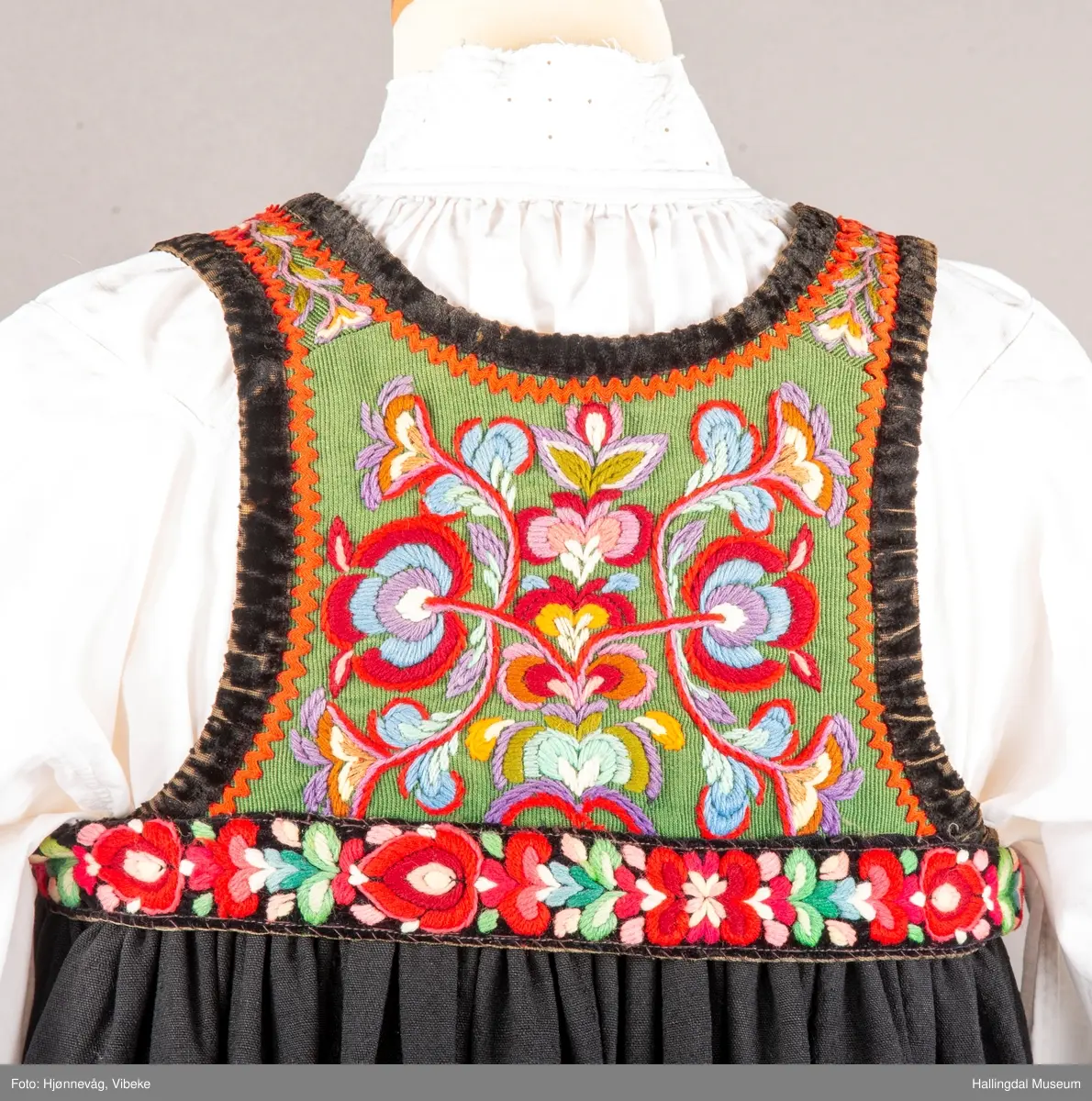 Ull, toskaft, sydd sammen av 5 høyder ca. 52 cm brede. Håndsydd. Broderi med ullgarn, mange ulike farger, akantusranke nederst. Blå kroklisse, grønn ullkant nederst. Lomme i høyre side. Blå skoning med hvite striper. Livet er grønt, ripsbinding. Svarte føyelskanter m/ rød kroklisse. Hvitt lerretsfor. Broderte hallingroser med ullgarn, sydd gjennom foret. Sølvspenner på hver side av livet med et kjede.
