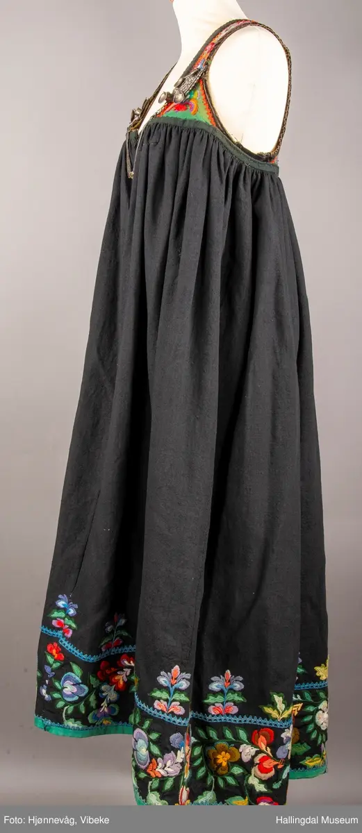 Ull, toskaft, sydd sammen av 5 høyder ca. 52 cm brede. Håndsydd. Broderi med ullgarn, mange ulike farger, akantusranke nederst. Blå kroklisse, grønn ullkant nederst. Lomme i høyre side. Blå skoning med hvite striper. Livet er grønt, ripsbinding. Svarte føyelskanter m/ rød kroklisse. Hvitt lerretsfor. Broderte hallingroser med ullgarn, sydd gjennom foret. Sølvspenner på hver side av livet med et kjede.