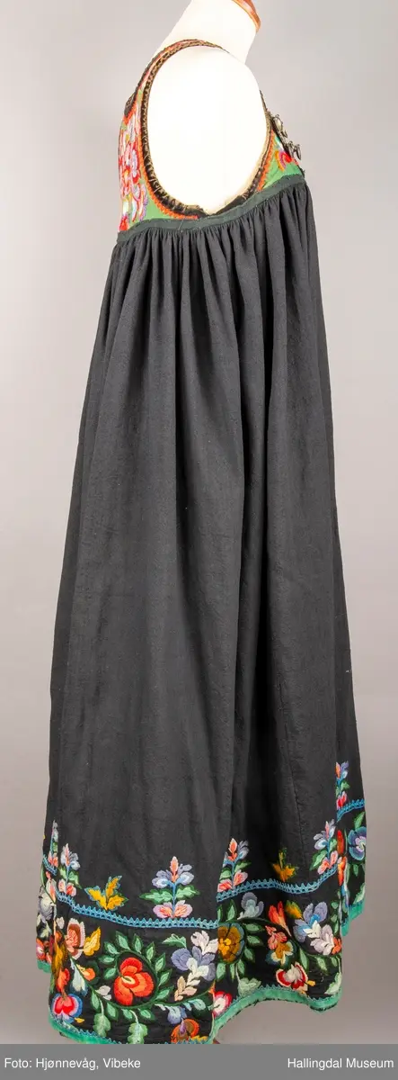 Ull, toskaft, sydd sammen av 5 høyder ca. 52 cm brede. Håndsydd. Broderi med ullgarn, mange ulike farger, akantusranke nederst. Blå kroklisse, grønn ullkant nederst. Lomme i høyre side. Blå skoning med hvite striper. Livet er grønt, ripsbinding. Svarte føyelskanter m/ rød kroklisse. Hvitt lerretsfor. Broderte hallingroser med ullgarn, sydd gjennom foret. Sølvspenner på hver side av livet med et kjede.