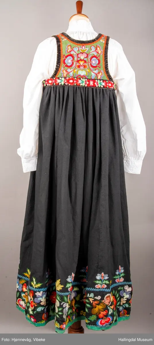 Ull, toskaft, sydd sammen av 5 høyder ca. 52 cm brede. Håndsydd. Broderi med ullgarn, mange ulike farger, akantusranke nederst. Blå kroklisse, grønn ullkant nederst. Lomme i høyre side. Blå skoning med hvite striper. Livet er grønt, ripsbinding. Svarte føyelskanter m/ rød kroklisse. Hvitt lerretsfor. Broderte hallingroser med ullgarn, sydd gjennom foret. Sølvspenner på hver side av livet med et kjede.