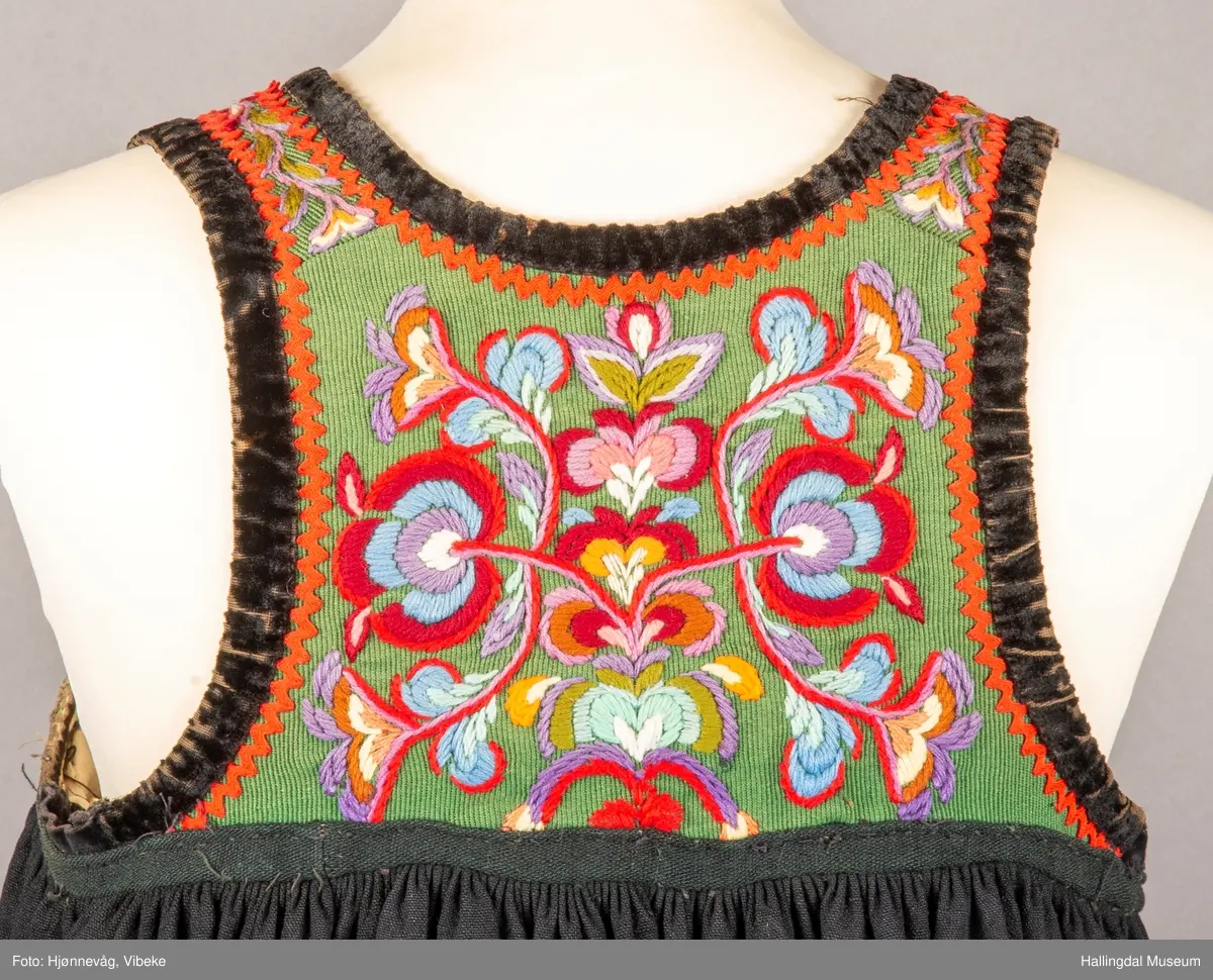 Ull, toskaft, sydd sammen av 5 høyder ca. 52 cm brede. Håndsydd. Broderi med ullgarn, mange ulike farger, akantusranke nederst. Blå kroklisse, grønn ullkant nederst. Lomme i høyre side. Blå skoning med hvite striper. Livet er grønt, ripsbinding. Svarte føyelskanter m/ rød kroklisse. Hvitt lerretsfor. Broderte hallingroser med ullgarn, sydd gjennom foret. Sølvspenner på hver side av livet med et kjede.