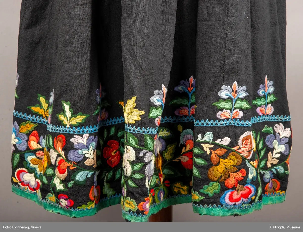 Ull, toskaft, sydd sammen av 5 høyder ca. 52 cm brede. Håndsydd. Broderi med ullgarn, mange ulike farger, akantusranke nederst. Blå kroklisse, grønn ullkant nederst. Lomme i høyre side. Blå skoning med hvite striper. Livet er grønt, ripsbinding. Svarte føyelskanter m/ rød kroklisse. Hvitt lerretsfor. Broderte hallingroser med ullgarn, sydd gjennom foret. Sølvspenner på hver side av livet med et kjede.