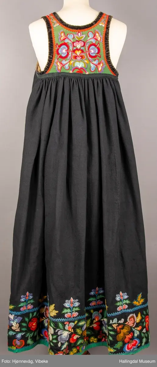 Ull, toskaft, sydd sammen av 5 høyder ca. 52 cm brede. Håndsydd. Broderi med ullgarn, mange ulike farger, akantusranke nederst. Blå kroklisse, grønn ullkant nederst. Lomme i høyre side. Blå skoning med hvite striper. Livet er grønt, ripsbinding. Svarte føyelskanter m/ rød kroklisse. Hvitt lerretsfor. Broderte hallingroser med ullgarn, sydd gjennom foret. Sølvspenner på hver side av livet med et kjede.