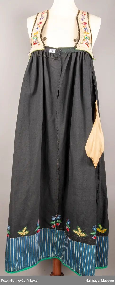 Ull, toskaft, sydd sammen av 5 høyder ca. 52 cm brede. Håndsydd. Broderi med ullgarn, mange ulike farger, akantusranke nederst. Blå kroklisse, grønn ullkant nederst. Lomme i høyre side. Blå skoning med hvite striper. Livet er grønt, ripsbinding. Svarte føyelskanter m/ rød kroklisse. Hvitt lerretsfor. Broderte hallingroser med ullgarn, sydd gjennom foret. Sølvspenner på hver side av livet med et kjede.