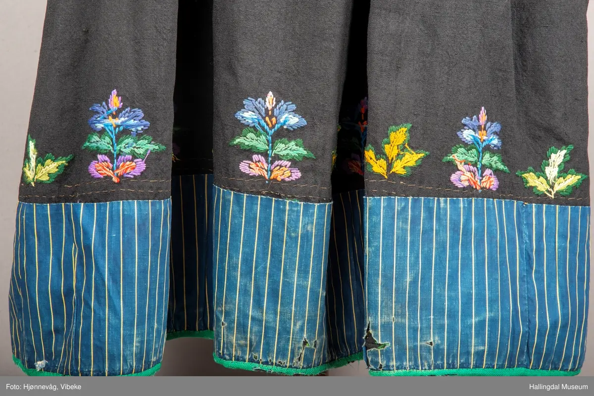 Ull, toskaft, sydd sammen av 5 høyder ca. 52 cm brede. Håndsydd. Broderi med ullgarn, mange ulike farger, akantusranke nederst. Blå kroklisse, grønn ullkant nederst. Lomme i høyre side. Blå skoning med hvite striper. Livet er grønt, ripsbinding. Svarte føyelskanter m/ rød kroklisse. Hvitt lerretsfor. Broderte hallingroser med ullgarn, sydd gjennom foret. Sølvspenner på hver side av livet med et kjede.
