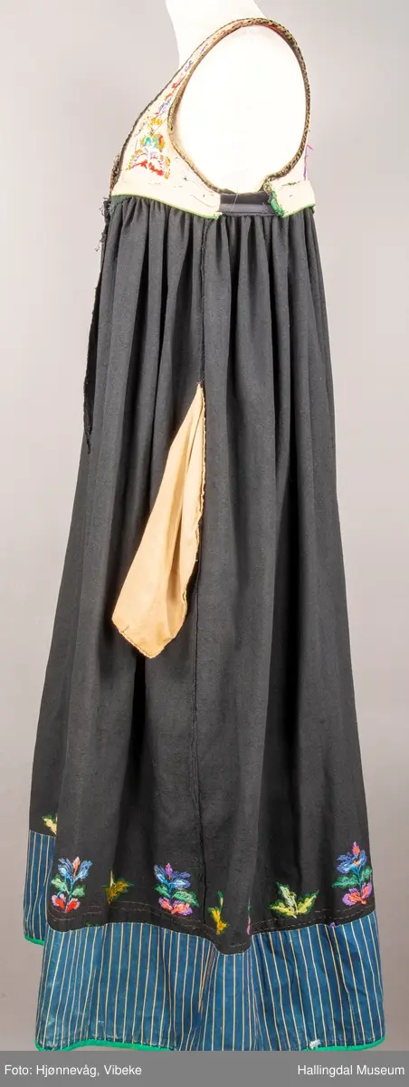 Ull, toskaft, sydd sammen av 5 høyder ca. 52 cm brede. Håndsydd. Broderi med ullgarn, mange ulike farger, akantusranke nederst. Blå kroklisse, grønn ullkant nederst. Lomme i høyre side. Blå skoning med hvite striper. Livet er grønt, ripsbinding. Svarte føyelskanter m/ rød kroklisse. Hvitt lerretsfor. Broderte hallingroser med ullgarn, sydd gjennom foret. Sølvspenner på hver side av livet med et kjede.