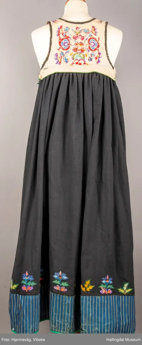 Ull, toskaft, sydd sammen av 5 høyder ca. 52 cm brede. Håndsydd. Broderi med ullgarn, mange ulike farger, akantusranke nederst. Blå kroklisse, grønn ullkant nederst. Lomme i høyre side. Blå skoning med hvite striper. Livet er grønt, ripsbinding. Svarte føyelskanter m/ rød kroklisse. Hvitt lerretsfor. Broderte hallingroser med ullgarn, sydd gjennom foret. Sølvspenner på hver side av livet med et kjede.