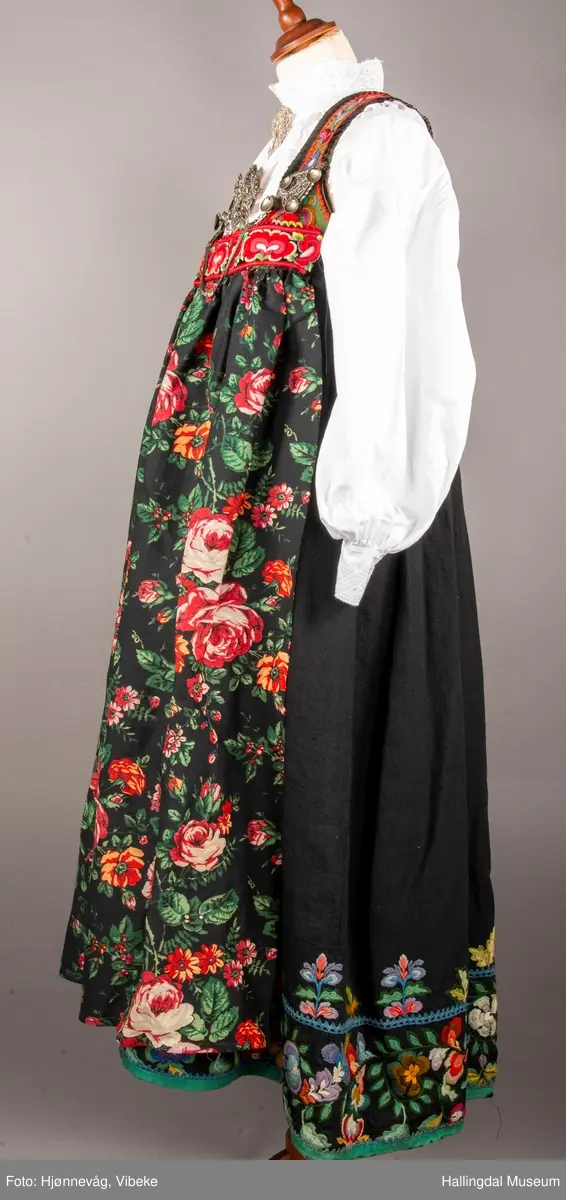 Ull, toskaft, sydd sammen av 5 høyder ca. 52 cm brede. Håndsydd. Broderi med ullgarn, mange ulike farger, akantusranke nederst. Blå kroklisse, grønn ullkant nederst. Lomme i høyre side. Blå skoning med hvite striper. Livet er grønt, ripsbinding. Svarte føyelskanter m/ rød kroklisse. Hvitt lerretsfor. Broderte hallingroser med ullgarn, sydd gjennom foret. Sølvspenner på hver side av livet med et kjede.