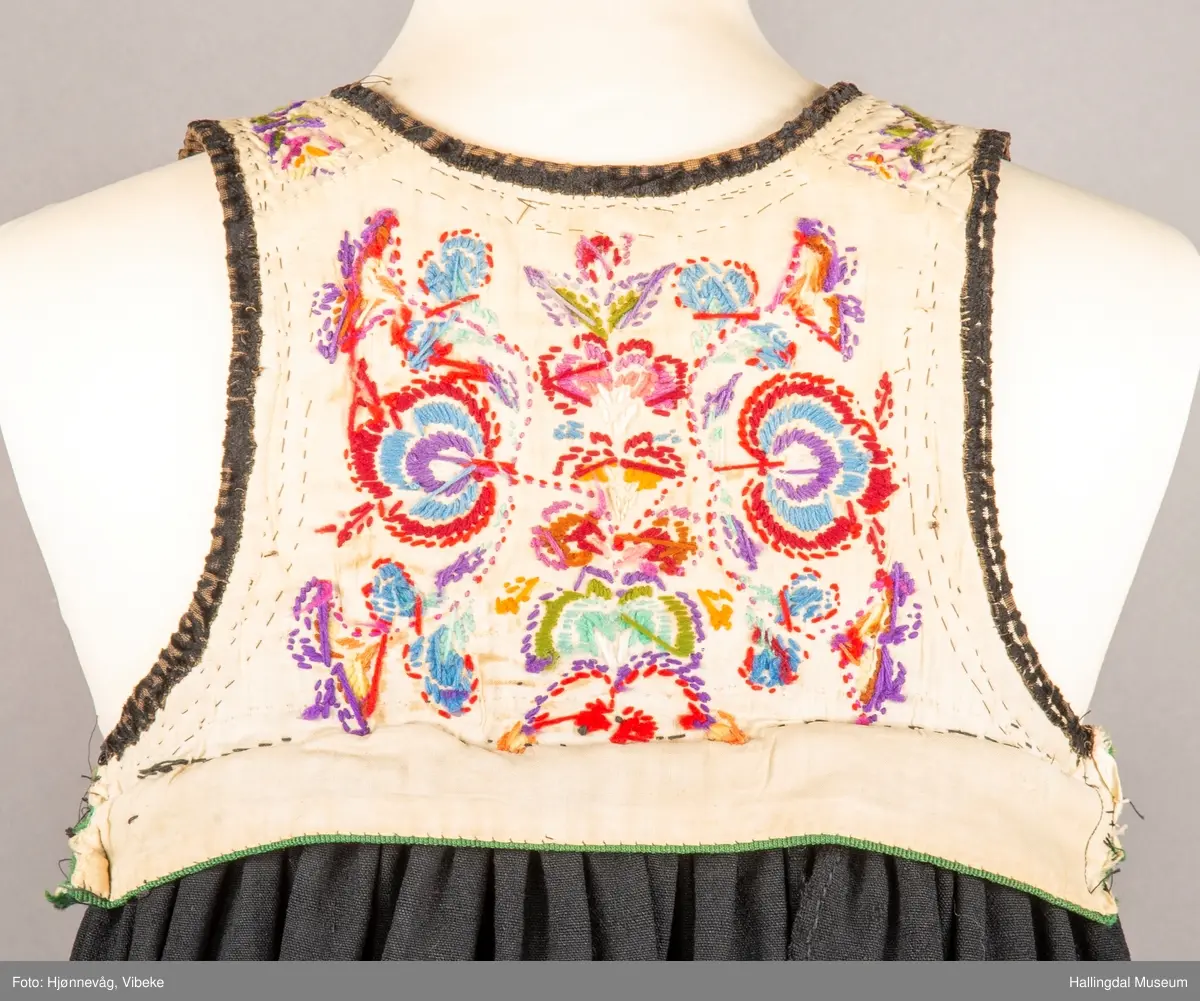 Ull, toskaft, sydd sammen av 5 høyder ca. 52 cm brede. Håndsydd. Broderi med ullgarn, mange ulike farger, akantusranke nederst. Blå kroklisse, grønn ullkant nederst. Lomme i høyre side. Blå skoning med hvite striper. Livet er grønt, ripsbinding. Svarte føyelskanter m/ rød kroklisse. Hvitt lerretsfor. Broderte hallingroser med ullgarn, sydd gjennom foret. Sølvspenner på hver side av livet med et kjede.