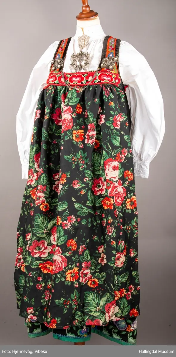 Ull, toskaft, sydd sammen av 5 høyder ca. 52 cm brede. Håndsydd. Broderi med ullgarn, mange ulike farger, akantusranke nederst. Blå kroklisse, grønn ullkant nederst. Lomme i høyre side. Blå skoning med hvite striper. Livet er grønt, ripsbinding. Svarte føyelskanter m/ rød kroklisse. Hvitt lerretsfor. Broderte hallingroser med ullgarn, sydd gjennom foret. Sølvspenner på hver side av livet med et kjede.