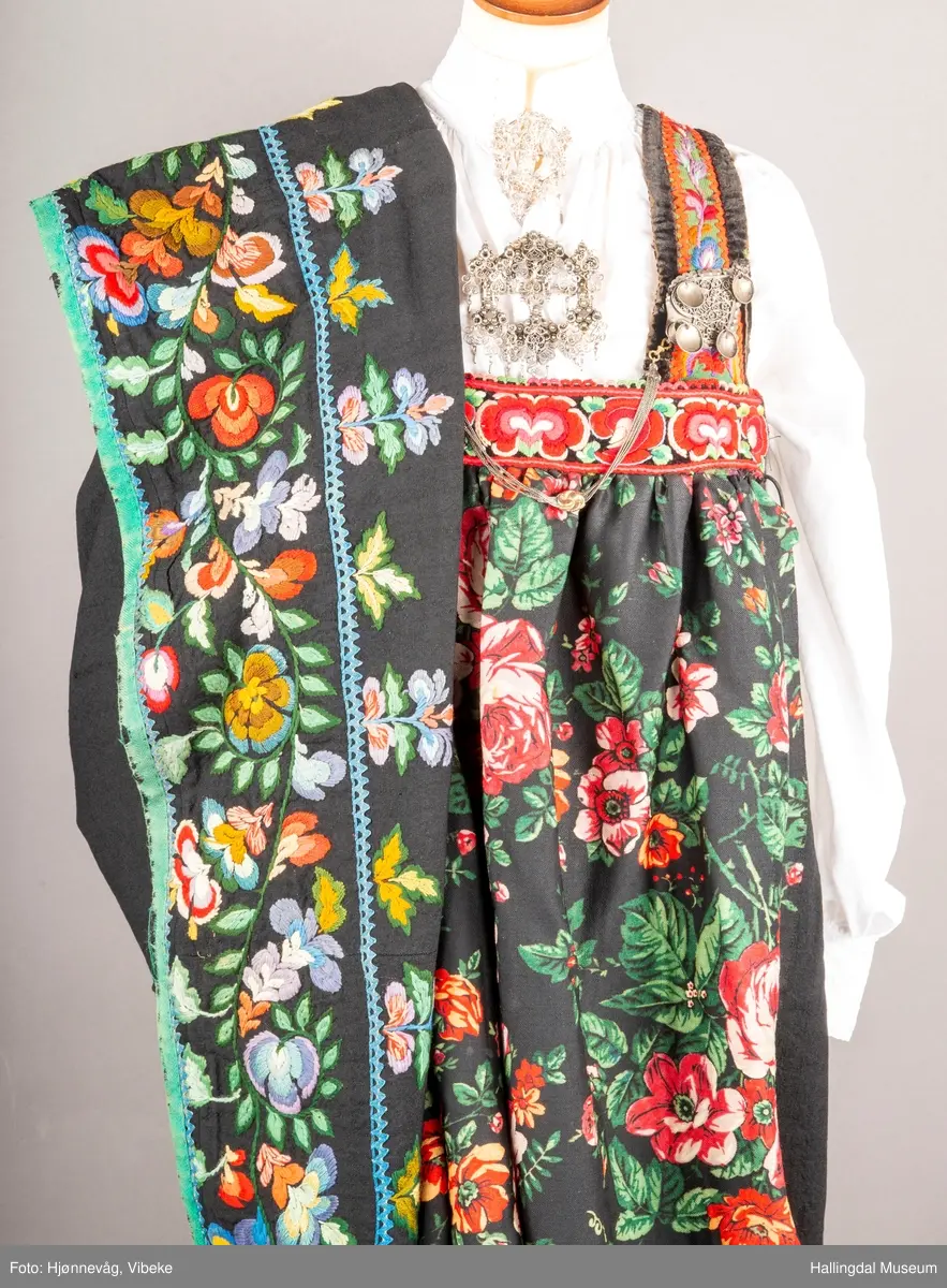 Ull, toskaft, sydd sammen av 5 høyder ca. 52 cm brede. Håndsydd. Broderi med ullgarn, mange ulike farger, akantusranke nederst. Blå kroklisse, grønn ullkant nederst. Lomme i høyre side. Blå skoning med hvite striper. Livet er grønt, ripsbinding. Svarte føyelskanter m/ rød kroklisse. Hvitt lerretsfor. Broderte hallingroser med ullgarn, sydd gjennom foret. Sølvspenner på hver side av livet med et kjede.