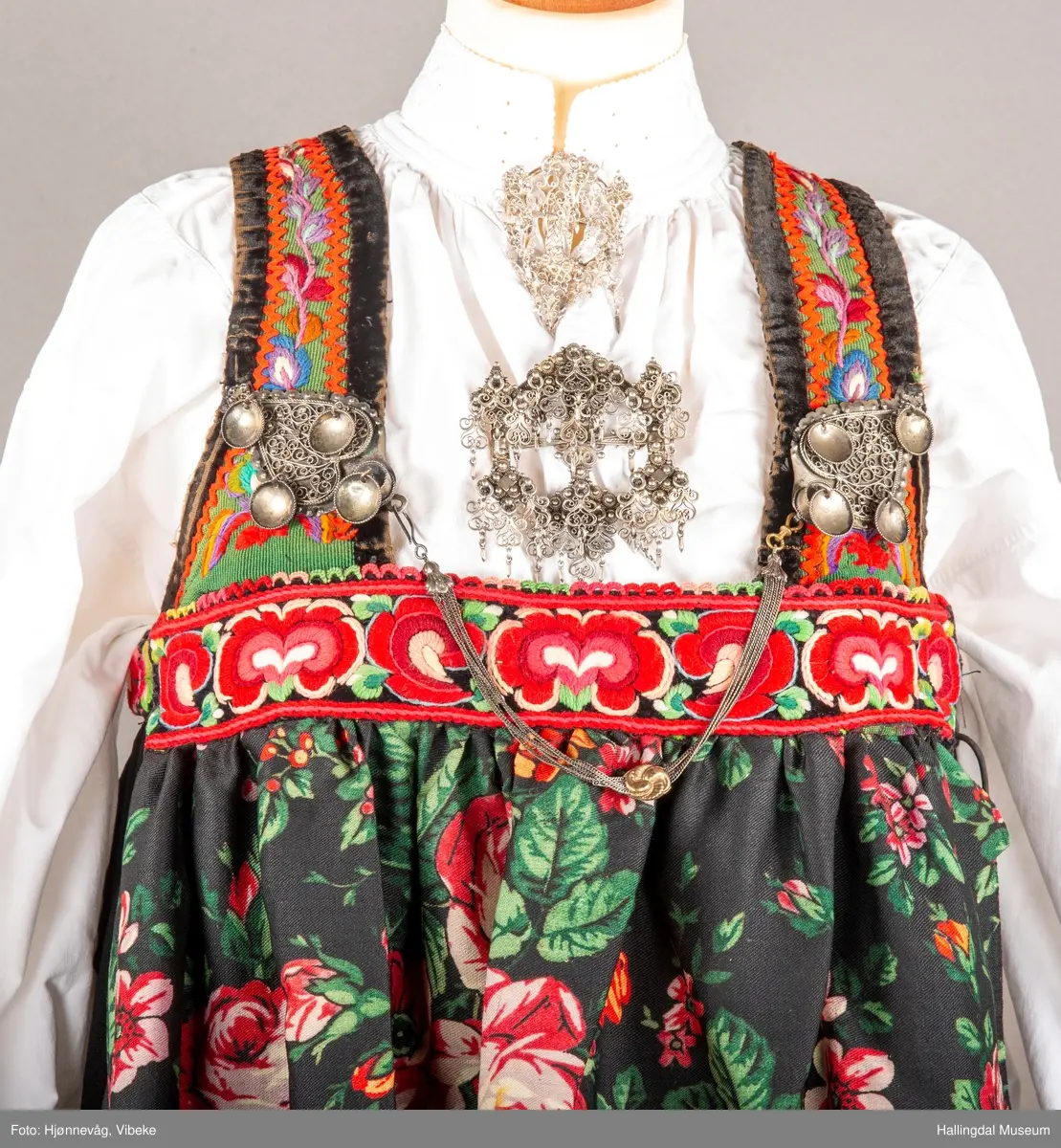 Ull, toskaft, sydd sammen av 5 høyder ca. 52 cm brede. Håndsydd. Broderi med ullgarn, mange ulike farger, akantusranke nederst. Blå kroklisse, grønn ullkant nederst. Lomme i høyre side. Blå skoning med hvite striper. Livet er grønt, ripsbinding. Svarte føyelskanter m/ rød kroklisse. Hvitt lerretsfor. Broderte hallingroser med ullgarn, sydd gjennom foret. Sølvspenner på hver side av livet med et kjede.