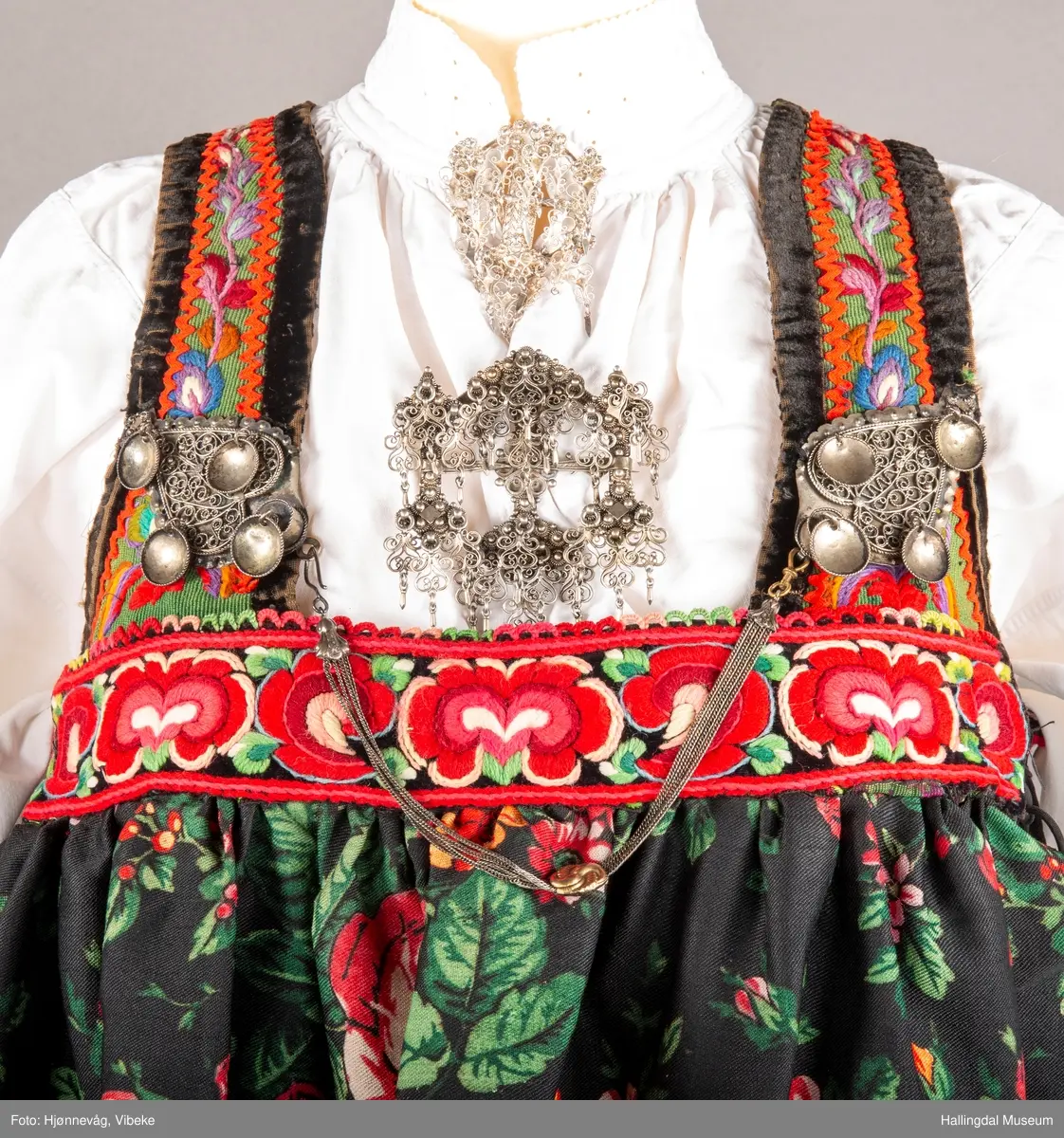 Ull, toskaft, sydd sammen av 5 høyder ca. 52 cm brede. Håndsydd. Broderi med ullgarn, mange ulike farger, akantusranke nederst. Blå kroklisse, grønn ullkant nederst. Lomme i høyre side. Blå skoning med hvite striper. Livet er grønt, ripsbinding. Svarte føyelskanter m/ rød kroklisse. Hvitt lerretsfor. Broderte hallingroser med ullgarn, sydd gjennom foret. Sølvspenner på hver side av livet med et kjede.