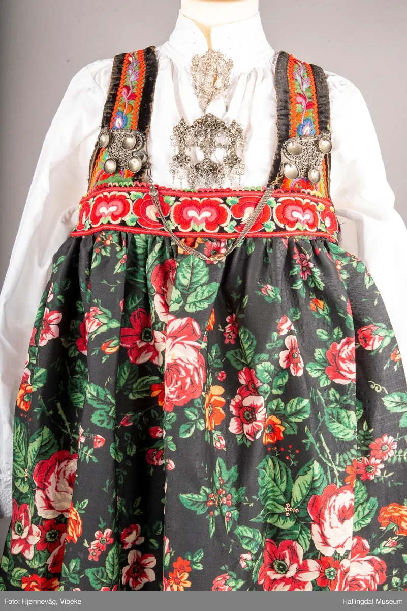 Ull, toskaft, sydd sammen av 5 høyder ca. 52 cm brede. Håndsydd. Broderi med ullgarn, mange ulike farger, akantusranke nederst. Blå kroklisse, grønn ullkant nederst. Lomme i høyre side. Blå skoning med hvite striper. Livet er grønt, ripsbinding. Svarte føyelskanter m/ rød kroklisse. Hvitt lerretsfor. Broderte hallingroser med ullgarn, sydd gjennom foret. Sølvspenner på hver side av livet med et kjede.