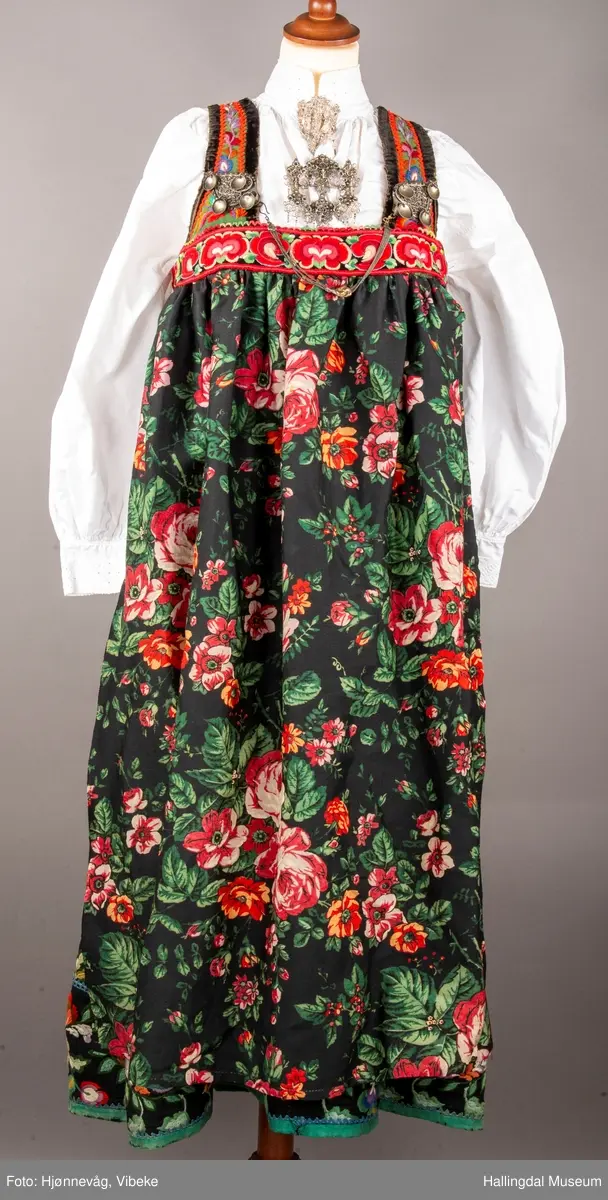 Ull, toskaft, sydd sammen av 5 høyder ca. 52 cm brede. Håndsydd. Broderi med ullgarn, mange ulike farger, akantusranke nederst. Blå kroklisse, grønn ullkant nederst. Lomme i høyre side. Blå skoning med hvite striper. Livet er grønt, ripsbinding. Svarte føyelskanter m/ rød kroklisse. Hvitt lerretsfor. Broderte hallingroser med ullgarn, sydd gjennom foret. Sølvspenner på hver side av livet med et kjede.