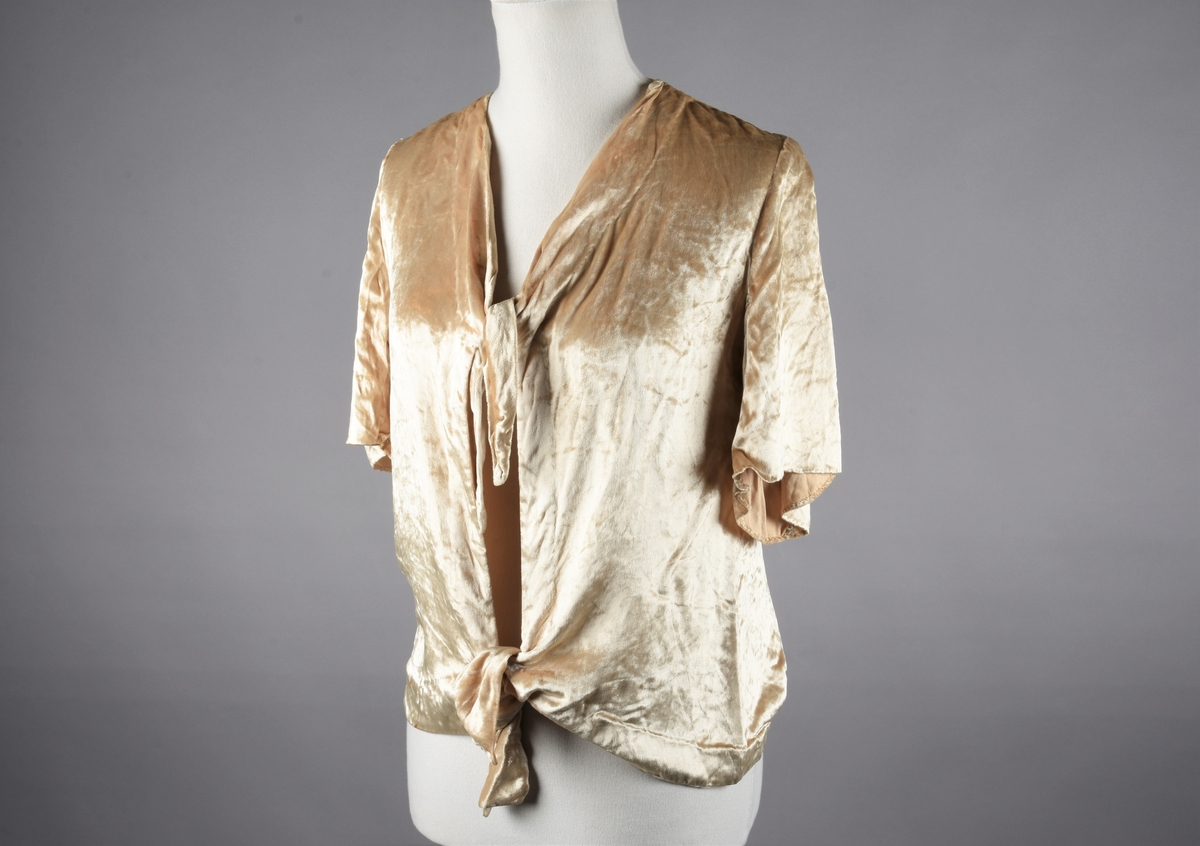 Elfenbensfarget bluse i tynn skinnende velour med flat lo. Alle kanter unntatt ermene har innvendig belegg av stoffet, og alle innvendige sømmer er kastet over med knapphullsting. De korte ermene er åpent traktformede. Eneste lukketøy er at korte brede knyttebånd er skåret i ett med forstykkene øverst og nederst. Halsåpningen er V-formet, og blusens nedre kant er skåret i bue oppover mot knytebåndene.