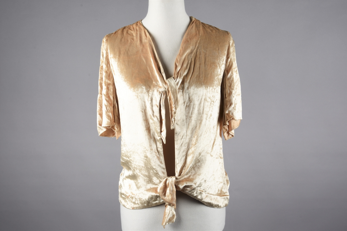 Elfenbensfarget bluse i tynn skinnende velour med flat lo. Alle kanter unntatt ermene har innvendig belegg av stoffet, og alle innvendige sømmer er kastet over med knapphullsting. De korte ermene er åpent traktformede. Eneste lukketøy er at korte brede knyttebånd er skåret i ett med forstykkene øverst og nederst. Halsåpningen er V-formet, og blusens nedre kant er skåret i bue oppover mot knytebåndene.
