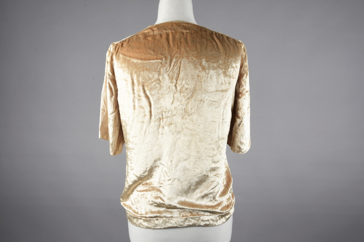 Elfenbensfarget bluse i tynn skinnende velour med flat lo. Alle kanter unntatt ermene har innvendig belegg av stoffet, og alle innvendige sømmer er kastet over med knapphullsting. De korte ermene er åpent traktformede. Eneste lukketøy er at korte brede knyttebånd er skåret i ett med forstykkene øverst og nederst. Halsåpningen er V-formet, og blusens nedre kant er skåret i bue oppover mot knytebåndene.