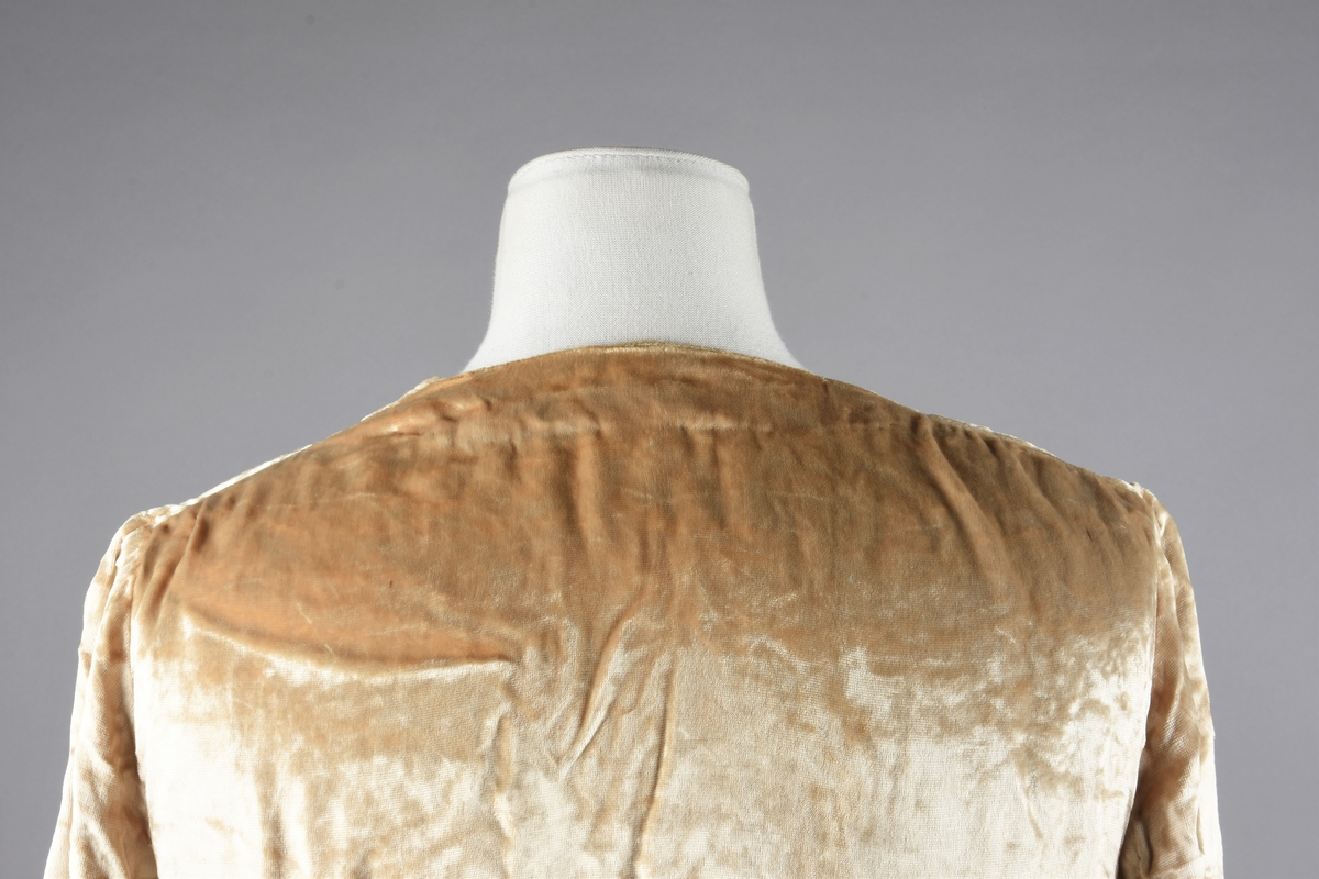 Elfenbensfarget bluse i tynn skinnende velour med flat lo. Alle kanter unntatt ermene har innvendig belegg av stoffet, og alle innvendige sømmer er kastet over med knapphullsting. De korte ermene er åpent traktformede. Eneste lukketøy er at korte brede knyttebånd er skåret i ett med forstykkene øverst og nederst. Halsåpningen er V-formet, og blusens nedre kant er skåret i bue oppover mot knytebåndene.