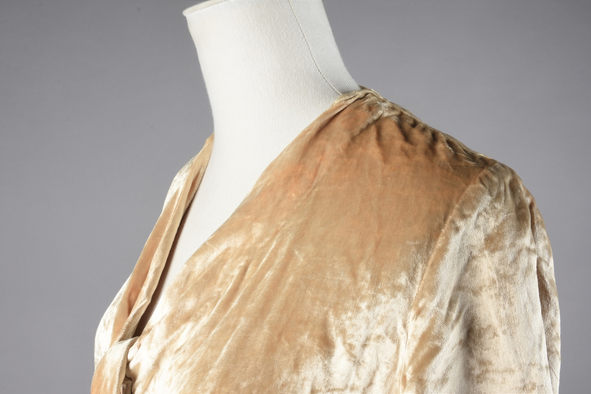 Elfenbensfarget bluse i tynn skinnende velour med flat lo. Alle kanter unntatt ermene har innvendig belegg av stoffet, og alle innvendige sømmer er kastet over med knapphullsting. De korte ermene er åpent traktformede. Eneste lukketøy er at korte brede knyttebånd er skåret i ett med forstykkene øverst og nederst. Halsåpningen er V-formet, og blusens nedre kant er skåret i bue oppover mot knytebåndene.