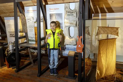 En liten gutt med helt ny og gul redningsvest poserer ved siden av en veldig gammel redningsvest som du finner i utstillingen det eventyrlige fisket