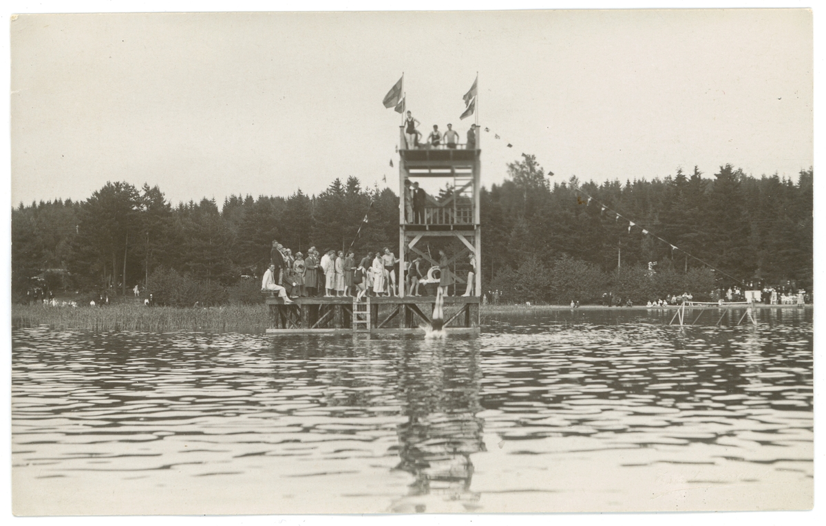 Motiv från Bredsand, Enköping. En samling människor på ett hopptorn. På kortets baksida står årtalet 1928. Vykortet ingår i ett album med vykort från Enköping (EM9535).