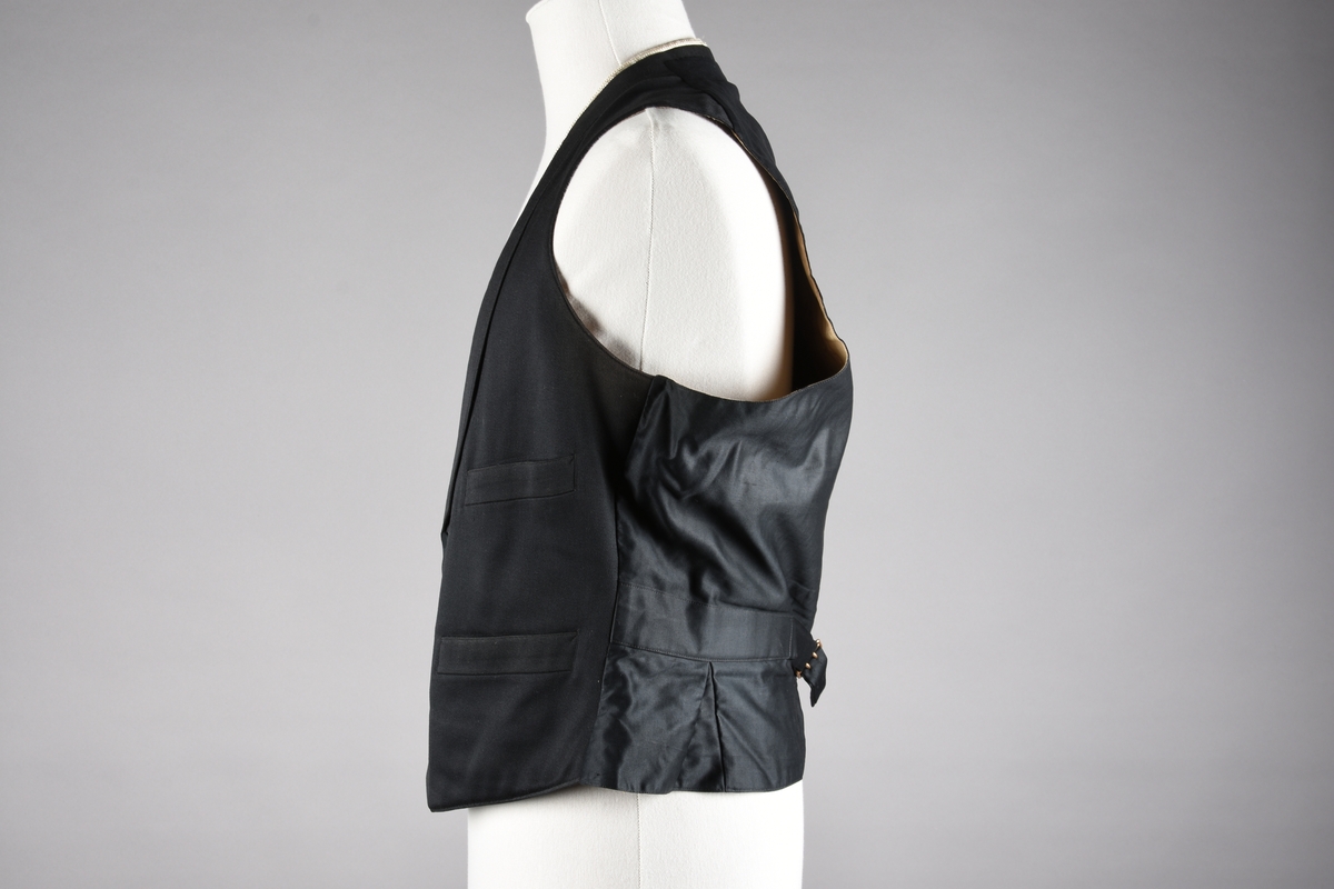 Vest av ull. God kvalitet på søm og materialer. Fôret med beige bomullssateng med lyseblå og røde striper, samt plantemotiv som er vevd inn i stoffet. Vesten lukkes med fire knapper som er trukket med samme stoff som vesten. Tre stikklommer. Tamp med metallspenne på ryggen. Kragen er kantet med hvitt possement. Possementet på vesten gir inntrykk av to vester på samme tid, altså med en «hvit vest» under. Dette To vester på denne måten ble ansett som veldig elegant.