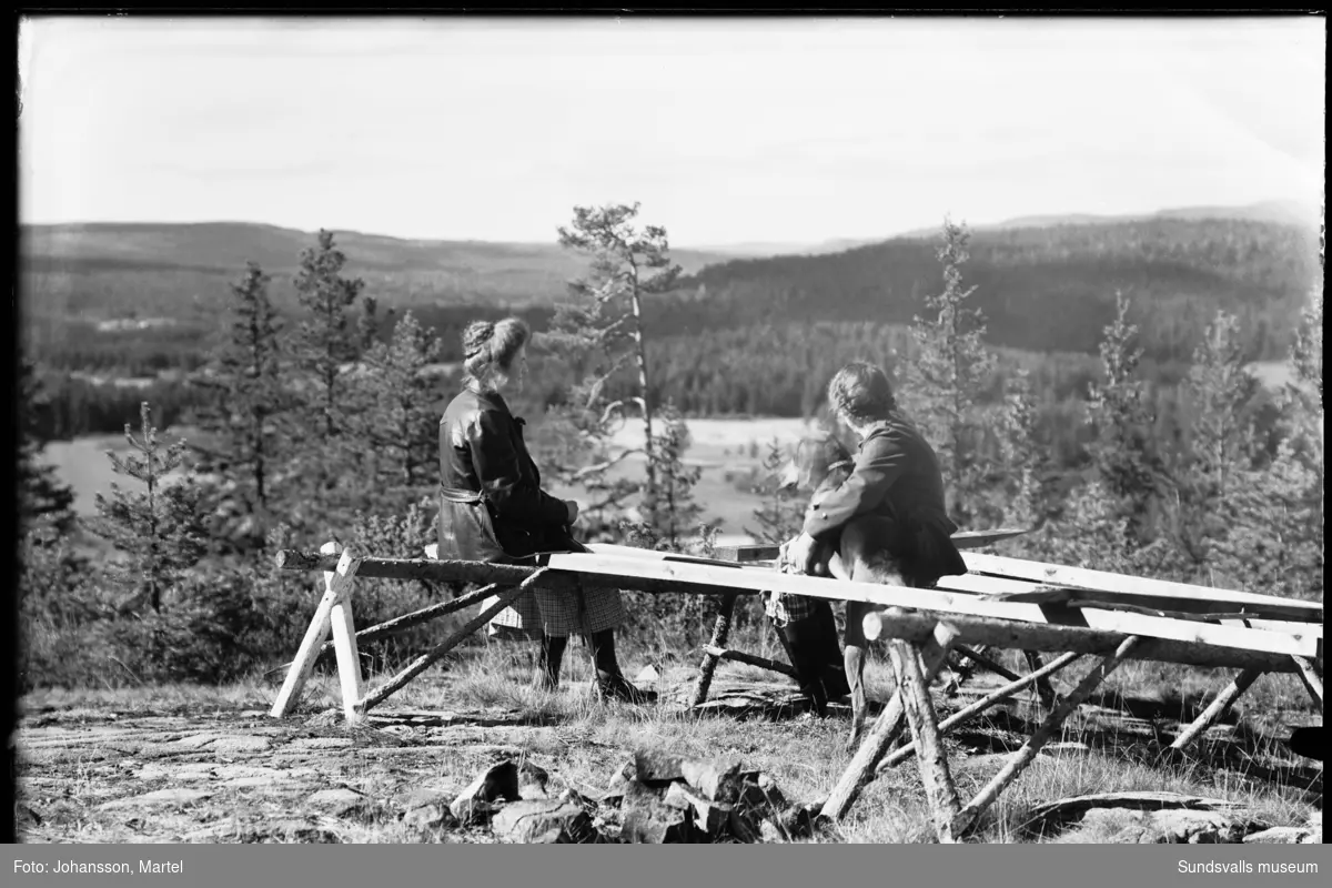 Två kvinnor sitter uppe på ett berg och tittar på utsikten. Den ena kvinnan har en stor hund i famnen. Möjligen befinner dom sig på Borgaråsen i Attmar.