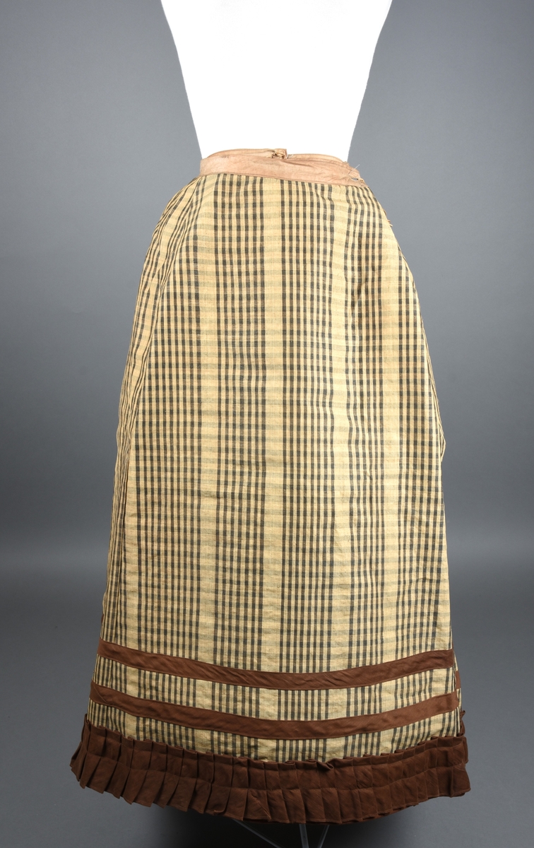 Stiveskjørt (underskjørt) av gul og gråbrunt-rutet nesleduk (?). Nederst er det en foldet kappe av gyldenbrun alpakka (?) med to rander over av skråstrimler av samme stoff som muligens er nyere. Skoning håndsydd av fint, ubleket lin. Antatt sekundær bruk fordi den er skjøtet sammen av veldig mange småstykker.