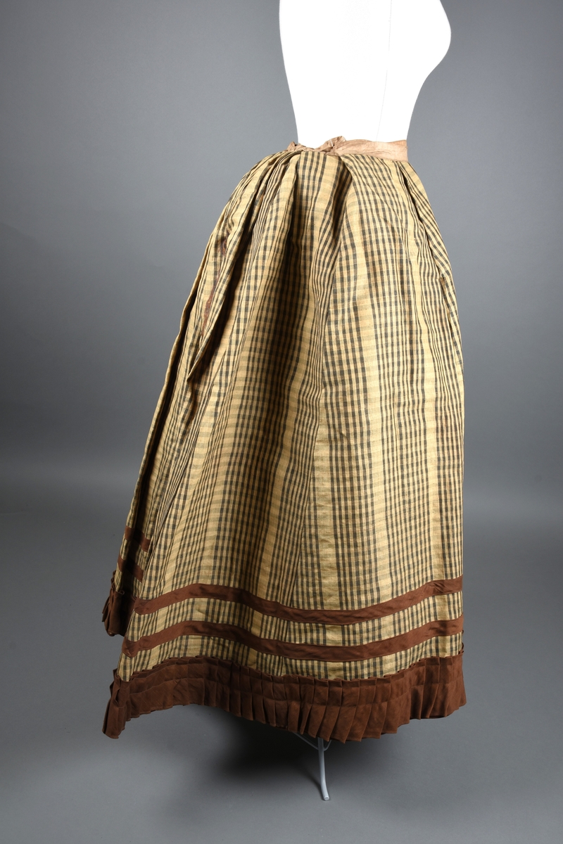 Stiveskjørt (underskjørt) av gul og gråbrunt-rutet nesleduk (?). Nederst er det en foldet kappe av gyldenbrun alpakka (?) med to rander over av skråstrimler av samme stoff som muligens er nyere. Skoning håndsydd av fint, ubleket lin. Antatt sekundær bruk fordi den er skjøtet sammen av veldig mange småstykker.