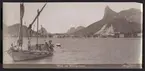 Bilden föreställer en vy över viken Baie de Botafogo i Rio de Janeiro från havet. På vänster sidan syns en segelbåt som transporterar en lass tegelpannor.