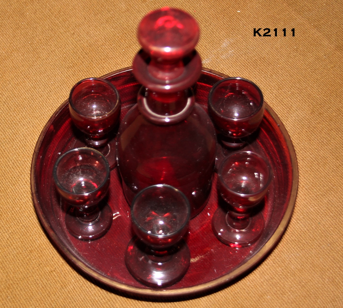 K2ll1, a—h. 
Likørkaraffel med 6 glass på brett, 1850—årene. Rubinglass, bøhmisk. 
a) Brett, sirkulært med skrå karm og forgylt kant.
b) Karaffel.
Runde skuldre, en ring om den smale hals, munningsbrem og propp. 
e-h) Glass, 6 stk. Rund fot, balusterstett, klokkeformet cupa.