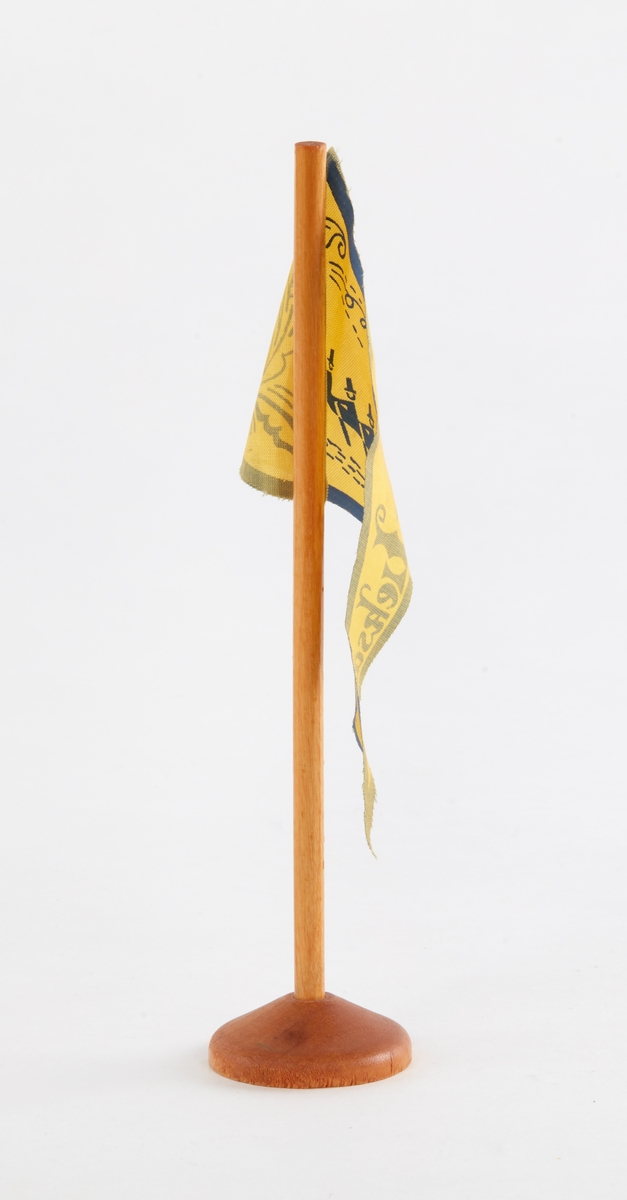 Bordsflagga, souvenir. Trekantig, gul flagga med blått tryck: 'Leksand', kyrkbåtsrodd och kurbits.